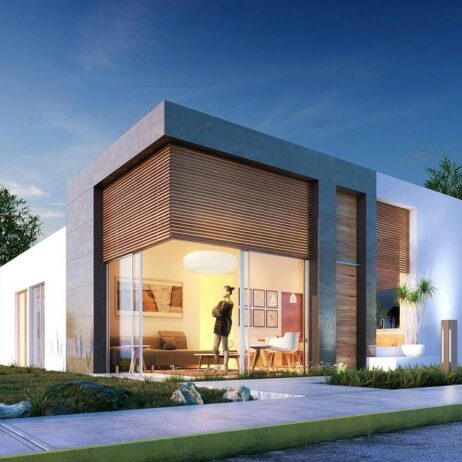 Vista exterior de los planos de casa pequeña Mía de una planta al atardecer, mostrando la fachada blanca y un volumen de concreto oscuro sobre el ventanal principal. La luz interior resalta el espacio de la sala y comedor.