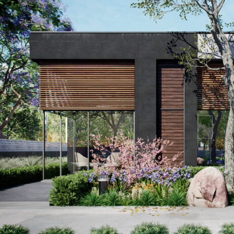 Vista frontal de [Planos de Casa pequeña Mía] casa de una planta con fachada oscura, persianas de madera y grandes ventanales. Se aprecian jardines floridos y árboles.