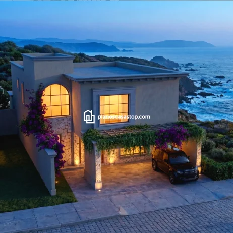 Planos de casa colonial Sauce: Elegancia y Vista al Mar Fachada de los Planos de casa colonial Sauce frente al mar con muros de piedra, ventanas en arco y pérgola con flores.